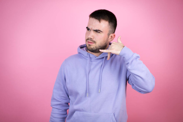 Jeune bel homme portant un sweat-shirt décontracté sur fond rose isolé confus faisant geste téléphonique avec la main et les doigts comme parler au téléphone - Photo, image