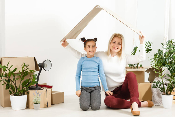 Cute mother and little daughter are on floor near boxes. They are preparing to move in another building. The parent and child are relaxing and smiling - Foto, Bild