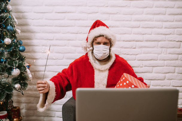 ビデオ通話上のラップトップの前でスプリンクラーと赤いギフトボックスを保持しながら、マスクを持つ幸せな若いサンタクロース。COVID中の新年とクリスマス19コロナウイルス - 写真・画像