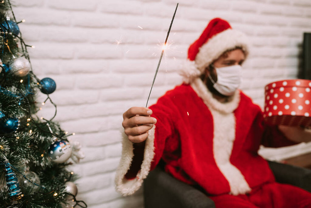 Papai Noel com um chapéu e máscara facial sentado na frente de um laptop e segurando aspersor e caixa de presente vermelho ao lado da árvore de Natal. Celebração do Ano Novo e Natal em quarentena COVID - 19 - Foto, Imagem