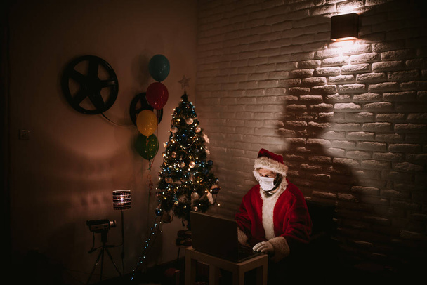 ラップトップの前に座ってクリスマスツリーの横のビデオ通話で話している帽子と顔のマスクを持つサンタクロース。隔離されたCOVIDの新年とクリスマスのお祝い- 19 - 写真・画像