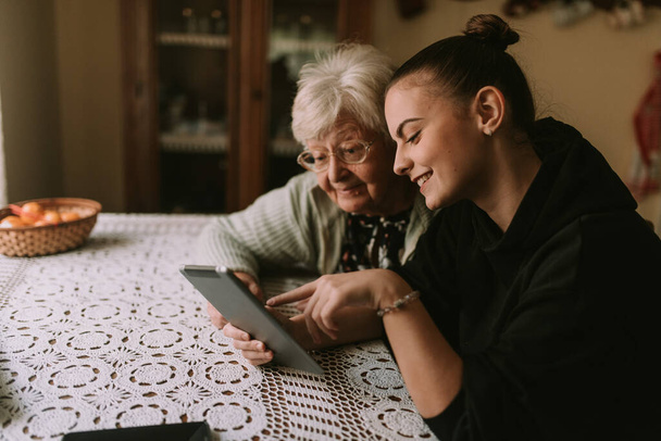 Roztomilá šedovlasá babička v důchodu a její dospívající vnučka se dívají na tabulku, jak sedí u stolu v babiččině domě. Babiččino vzdělání o nových technologiích - Fotografie, Obrázek