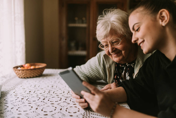 A smiling caucasian grandmother and her teenage granddaughter look at a tablet while sitting at a table in the kitchen. The granddaughter teaches her grandmother to use new technology - Photo, Image