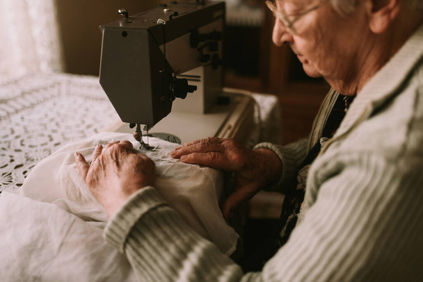 Close-up de uma mulher caucasiana sênior com óculos costurando uma cortina em uma máquina de costura em uma mesa na casa. Passatempos prediletos de mulheres seniores - Foto, Imagem