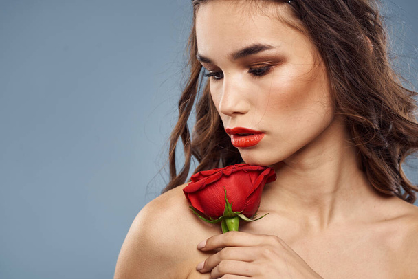 Γυναίκα πορτρέτο με κόκκινο τριαντάφυλλο κοντά στο πρόσωπο σε γκρι φόντο και μακιγιάζ σγουρά μαλλιά - Φωτογραφία, εικόνα