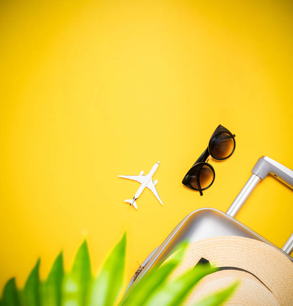 Тропический фон. Чемодан, солнцезащитные очки с пальмовыми листьями и соломенной шляпой, белая плоскость в композиции путешествия на желтом фоне. Экзотический тропический пляж с копировальным пространством - Фото, изображение