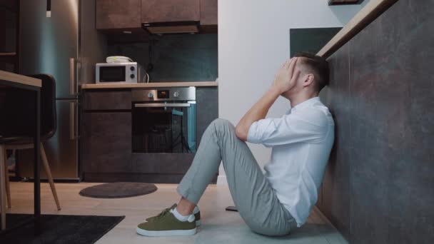 Młody człowiek w depresji siedzi na podłodze w kuchni. Zdesperowany biznesmen zakrywa twarz rękami i płacze.. - Materiał filmowy, wideo