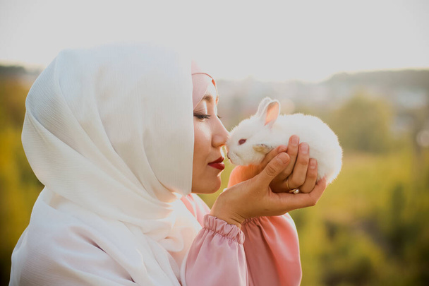 Muslim woman hold a small white rabbit in her arms. A portrait of a Muslim woman in a pink dress and a scarf, a pearl bracelet. Tenderness. - Foto, Imagen