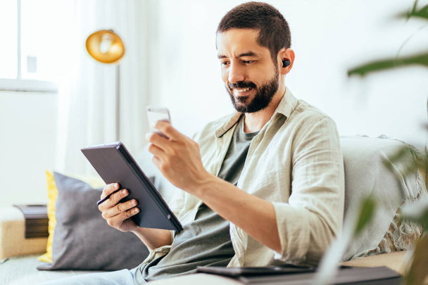 Young bearded man working from home. Man on sofa at home using digital tablet - 写真・画像