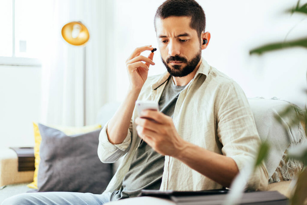 Hombre poniéndose auriculares para hacer videollamadas desde su celular en el sofá de su casa. - Foto, imagen