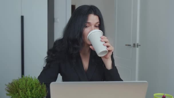Žena používá notebook, zatímco sedí u stolu a pije kávu. Mladá evropská podnikatelka sedí v kanceláři a pracuje na počítačovém softwaru. Záběr steadicamu. - Záběry, video