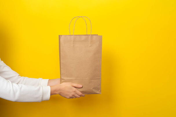 Female hands hold large gift bag made of brown craft paper on yellow background. Gift, present or e-shopping concept. Banner with copy space - Fotografie, Obrázek