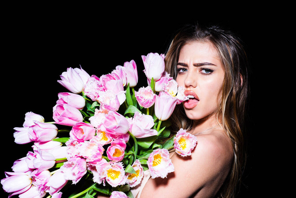 Young woman with flowers. Beautiful girl holding bouquet of tulips over black background sticking tongue with sexy expression. - Fotó, kép