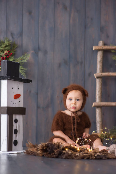 Happy little boy in a brown, fluffy, knitted bear costume, sits on the furon the wooden floor, plays with a bright Christmas garland of lights. There is a snowman nearby. New year card. Copy space  - Fotografie, Obrázek