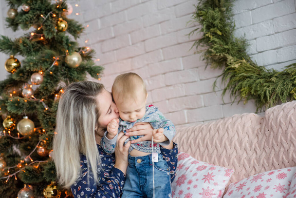 A beautiful young woman sits on a sofa with a boy near Christmas tree against a white brick wall. Mother hugs, kisses, plays with her small son. Happy family celebrates New Year. Copy space, festive  - Φωτογραφία, εικόνα