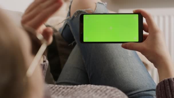 Giovane donna che tiene smartphone con schermo verde mentre riposa sul divano e agita. Videochiamata - Filmati, video