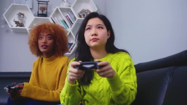 Iloinen onnellinen houkutteleva tytöt pelaavat videopelejä kotona. Keskittynyt aasialainen nainen menettää pelin afrikkalainen amerikkalainen musta ystävä - Materiaali, video