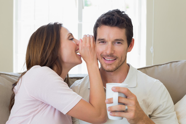 Woman whispering secret into a happy mans ear in living room - Фото, изображение