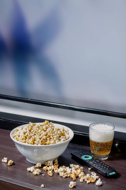 Eine Schüssel voll Popcorn, etwas Popcorn auf dem Tisch neben einem Pils vor einem LED-Fernseher mit Fernbedienung. - Foto, Bild