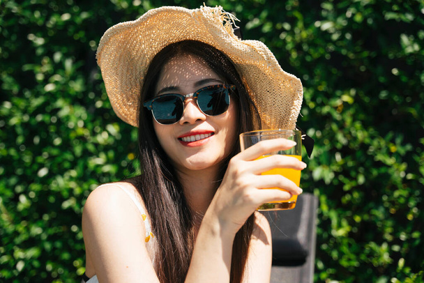 Retrato de viajero asiático mujer usar gafas de sol y sombrero disfrutar de jugo de naranja en verano. - Foto, Imagen