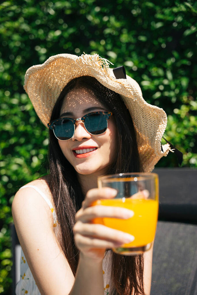 Πορτρέτο της ασιατικής ταξιδιώτη γυναίκα φορούν γυαλιά ηλίου και καπέλο απολαύσετε χυμό πορτοκάλι το καλοκαίρι. - Φωτογραφία, εικόνα