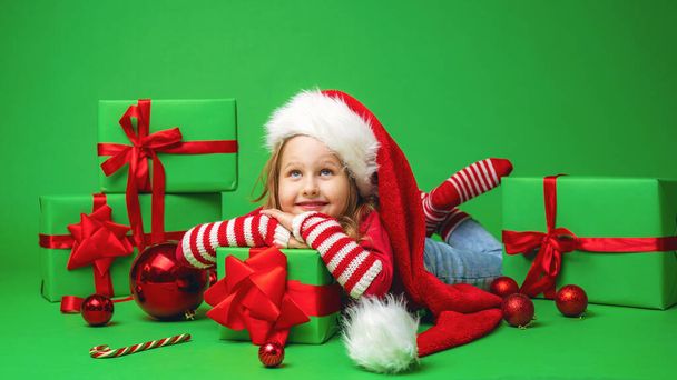 little girl in a Santa hat is lying on a gift box with a red bow on a green background in the Studio. the child smiles happily and looks up. Advertizing. Copy space. - Photo, image