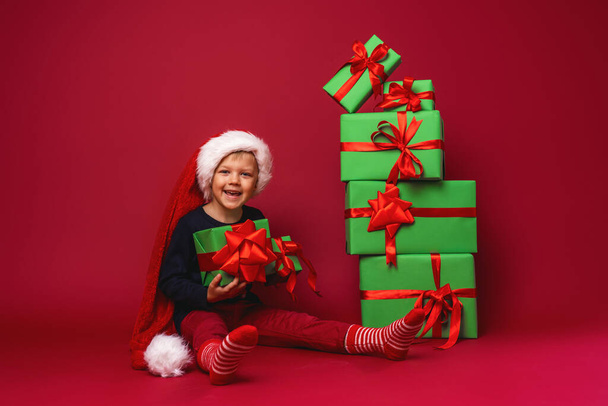 little boy in Santa hat sits next to pyramid Christmas gift boxes on red background in Studio. the child is happy to smile and hold gifts in his hands, get lot of gifts for Christmas. Advertizing. - 写真・画像