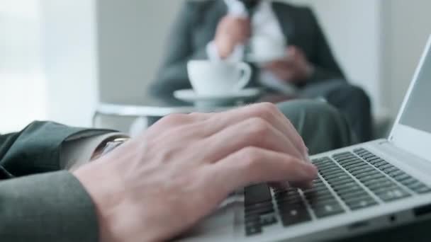 Unbekannter sitzt an niedrigem Tisch im Büroflur und tippt etwas auf Laptop, während er mit seinem Kollegen Kaffeepause macht - Filmmaterial, Video