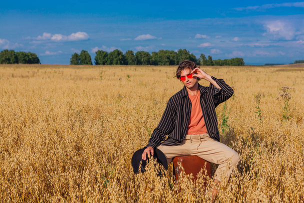 Rural Countryside Scene. Tall handsome man dressed in a black shirt, black hat and pink sunglasses sitting on a brown vintage leather suitcase at golden oat field. Summer landscape with blue sky - Fotoğraf, Görsel