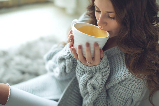 Close-up van vrouwelijke handen met een mok drank. Mooi meisje in een grijze trui houdt een kopje thee of koffie in de ochtend zonlicht. Mok voor uw ontwerp.  - Foto, afbeelding