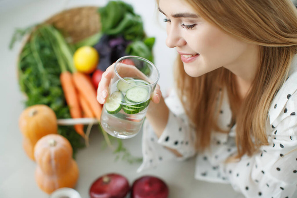 Young woman drinking water near the table with fruits and vegetables in the kitchen. - Photo, Image