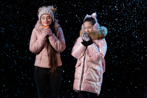 The frozen mother and daughter warm up and enjoy the aroma of the hot drink. Picture taken in a dark studio against a black background during a snowfall. Close up. - Foto, Bild