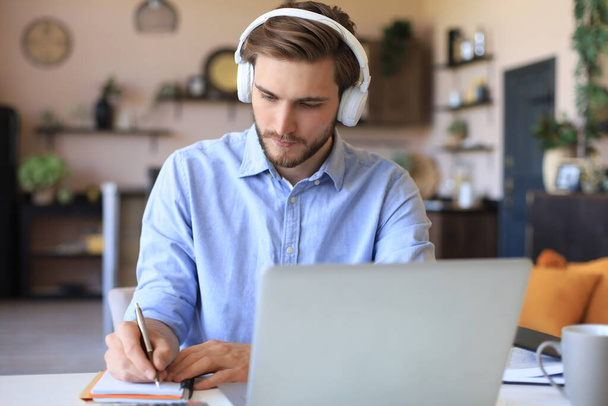 Confident businessman in earphones is writing notes or financial report while sitting at desk with laptop at home - Foto, imagen