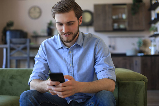 Attractive young man relaxing on a couch at home and using mobile phone for cheking social nets - Foto, Imagen