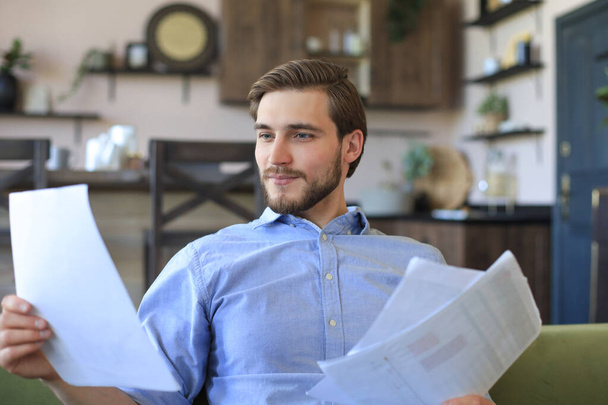 Concentrated young freelancer businessman sitting on sofa with laptop and examining documents - Foto, Imagem