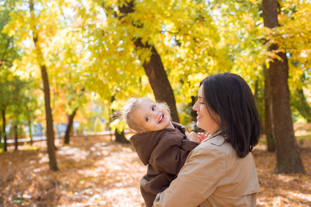 Μια νεαρή μητέρα και μια μικρή κόρη περπατούν και γελάνε στο φθινοπωρινό πάρκο. Οικογένεια. Φθινοπωρινή - Φωτογραφία, εικόνα