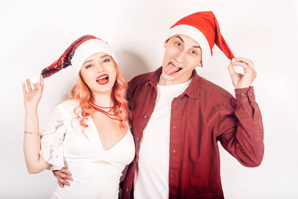Пара на белом фоне в шляпах Санты. Девушка и парень улыбаются, Новый год, Рождество. Женщина в красивом белом платье и розовые волосы, в аксессуарах. Любители гримасничают и веселятся - Фото, изображение