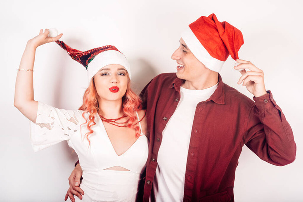 Paar auf weißem Hintergrund mit Weihnachtsmützen. Das Mädchen und der Kerl lächeln, Neujahr, Weihnachten. Frau in einem schönen weißen Kleid und rosa Haaren, in Accessoires. Liebhaber grimassieren und haben Spaß - Foto, Bild