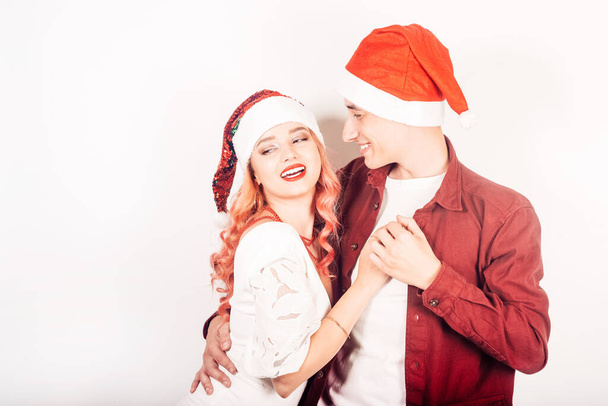 Couple on a white background in santa hats. The girl and the guy are smiling, new year, christmas. Woman in a beautiful white dress and pink hair, in accessories - 写真・画像
