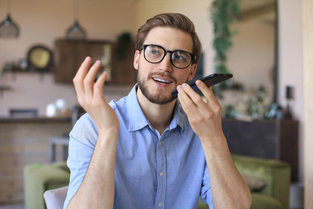 Confident business man in glasses holding smartphone near mouth for recording voice message or activating digital assistant - Photo, Image