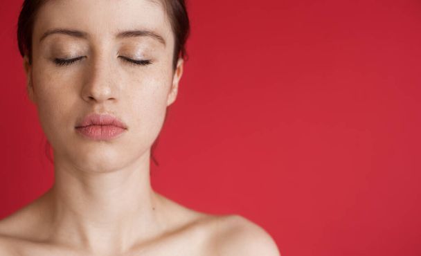 Λευκή γυναίκα με φακίδες ποζάρει με κλειστά μάτια και γυμνούς ώμους σε κόκκινο τοίχο στούντιο με ελεύθερο χώρο - Φωτογραφία, εικόνα