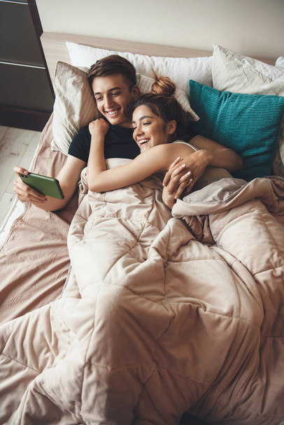 Φωτογραφία πάνω όψης ενός καυκάσιου ζευγαριού που κείτεται και αγκαλιάζεται στο κρεβάτι χαμογελώντας στο τηλέφωνο - Φωτογραφία, εικόνα