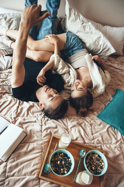 Foto vista superior de una pareja caucásica acostada en la cama y haciendo una selfie mientras come cereales con leche sonriendo al teléfono - Foto, imagen