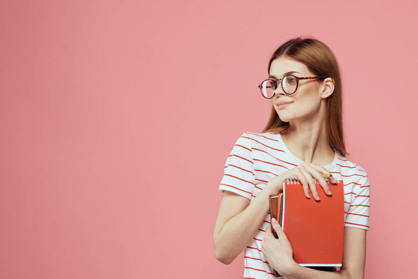 Nainen opiskelija muistilehtiöt vaaleanpunainen tausta elehtii kädet lasit kasvoillaan raidallinen t-paita malli - Valokuva, kuva