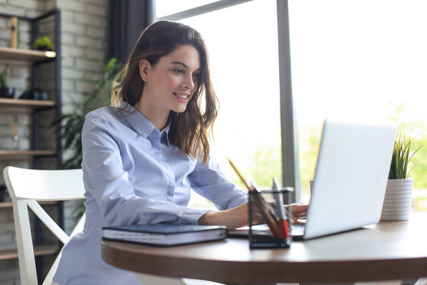 Smiling pretty woman sitting at table, looking at laptop screen. Happy entrepreneur reading message email with good news, chatting with clients online - Valokuva, kuva