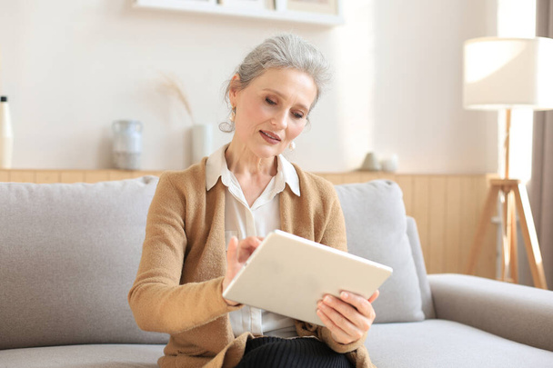 Mulher alegre de meia-idade sentado no sofá, usando aplicativos tablet computador, olhando para a tela, lendo boas notícias na rede social, compras ou bate-papo on-line - Foto, Imagem