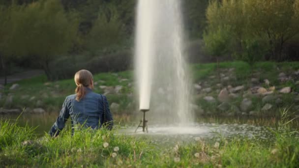 Eine junge Frau ruht in einem Park am Ufer eines kleinen Stausees. Sie blickt auf die Gischt des Brunnens und ist abgelenkt vom täglichen Trubel.. - Filmmaterial, Video