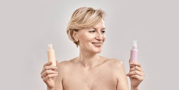 Portrait of beautiful middle aged woman holding two bottles of different cosmetic skincare products and choosing what to apply while posing isolated over grey background - Photo, image
