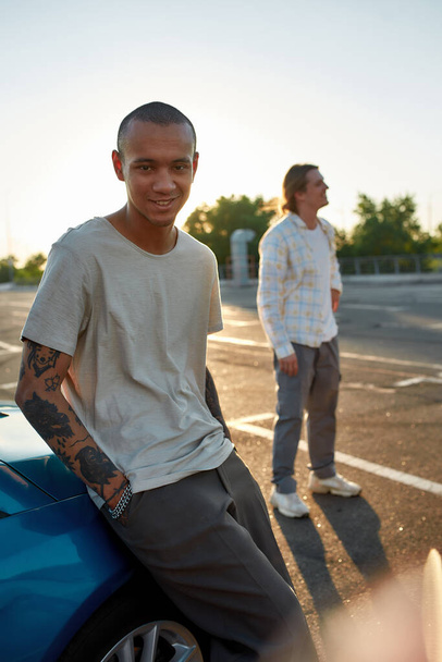 Egy sötét bőrű, tetovált fickó fehér ingben, zsebre tett kézzel, aki egy autó mellett áll egy parkolóban a barátaival a háttérben. - Fotó, kép