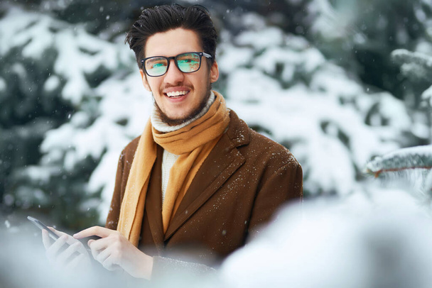 Young man with phone in winter park.  Businessman uses a phone and  enjoying winter moments in a snowy forest. Snowfall. - Zdjęcie, obraz
